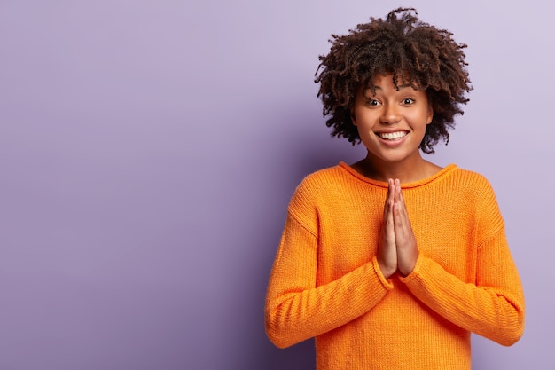 Sonriente mujer afroamericana ruega por algo, mantiene las palmas juntas en gesto de oración, tiene una mirada alegre suplicante