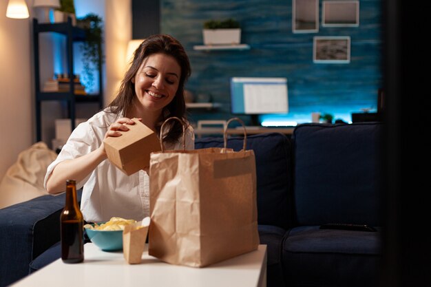 Sonriente mujer alegre desembalaje degustación de comida rápida a domicilio entregado sentado en el sofá