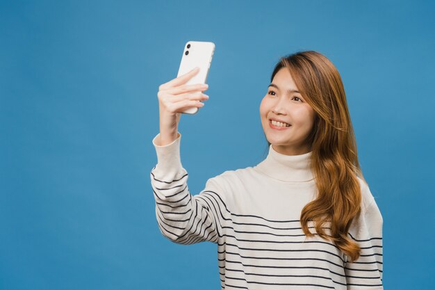 Sonriente mujer asiática adorable haciendo foto selfie en teléfono inteligente con expresión positiva en ropa casual y soporte aislado en la pared azul