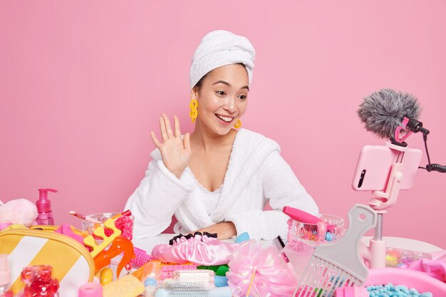 Sonriente mujer asiática belleza blogger películas maquillaje tutorial ondas hola en la cámara del teléfono inteligente