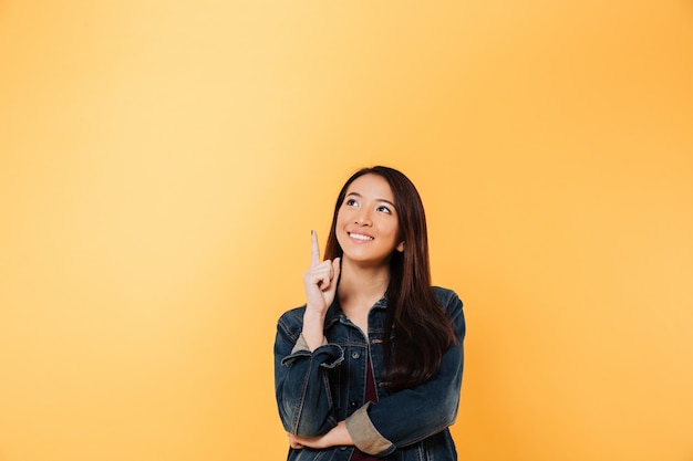 Sonriente mujer asiática en chaqueta vaquera apuntando y mirando hacia arriba sobre fondo amarillo