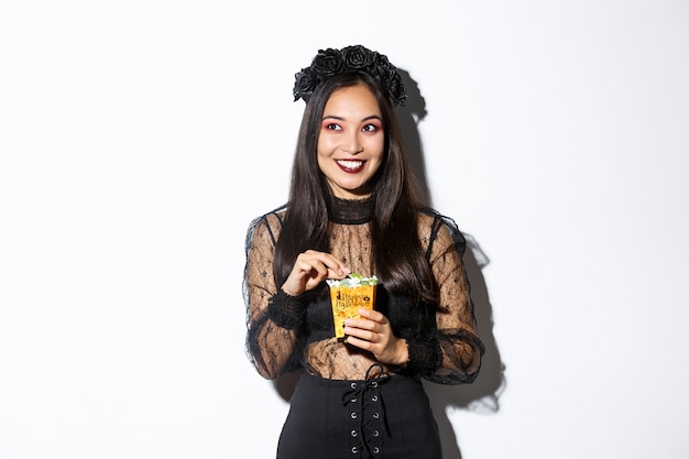 Sonriente mujer asiática linda celebrando halloween, sosteniendo dulces y sonriendo feliz, truco o trato en traje de bruja
