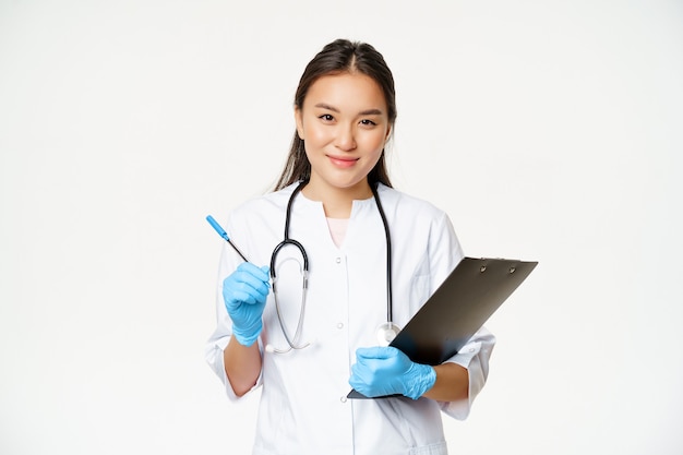 Sonriente mujer asiática médico enfermera sosteniendo portapapeles y bolígrafo vistiendo uniforme con guantes escribiendo pati ...