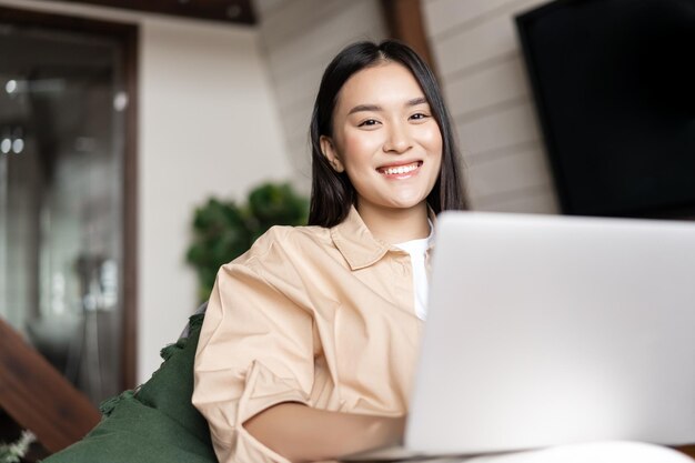 Sonriente mujer asiática con ordenador portátil en casa y mirando feliz a la cámara