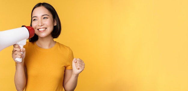 Sonriente mujer asiática de pie con megáfono anunciando algo de producto publicitario de pie sobre amarillo