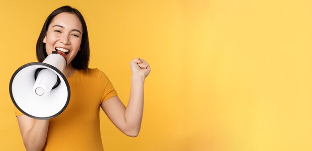 Sonriente mujer asiática de pie con megáfono anunciando algo de producto publicitario de pie sobre amarillo