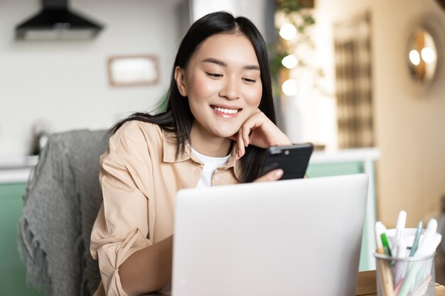 Sonriente mujer asiática que trabaja de forma remota desde casa usando una computadora portátil y un teléfono móvil sentado en la mesa con c ...