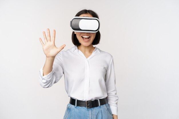 Sonriente mujer asiática saludándote con gafas de realidad virtual vr auriculares charlando en metauniverso de pie sobre fondo blanco.