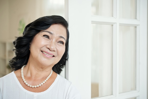 Sonriente mujer asiática senior recostada en la puerta de su casa y mirando a otro lado