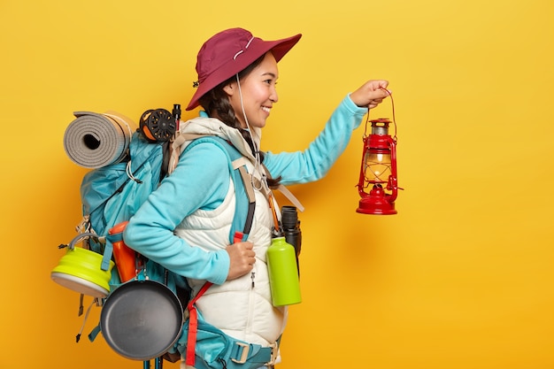 Sonriente mujer asiática sostiene una pequeña lámpara de aceite, va a explorar el lugar en la oscuridad, lleva una mochila con pertenencias personales