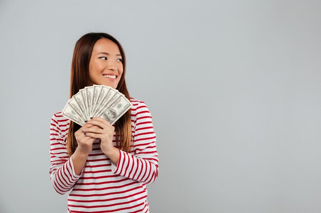 Sonriente mujer asiática en suéter con dinero y mirando a otro lado sobre fondo gris