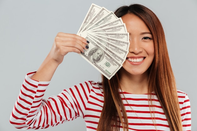 Sonriente mujer asiática en suéter está cubriendo en dinero de la mitad de la cara y mirando a la cámara sobre fondo gris