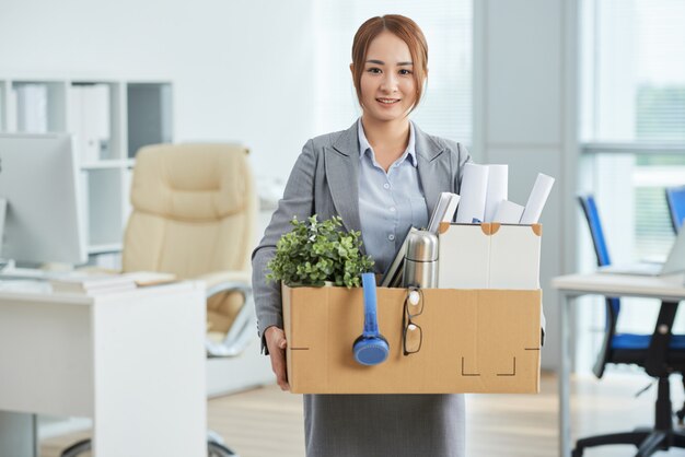 Sonriente mujer asiática en traje de negocios de pie en la oficina con sus pertenencias en caja de cartón