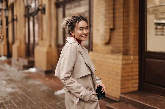 Sonriente mujer asiática vestida con abrigo beige de otoño sosteniendo bandolera y posando contra el edificio antiguo de ladrillo