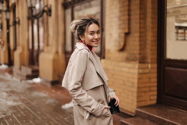 Sonriente mujer asiática vestida con abrigo beige de otoño sosteniendo bandolera y posando contra el edificio antiguo de ladrillo