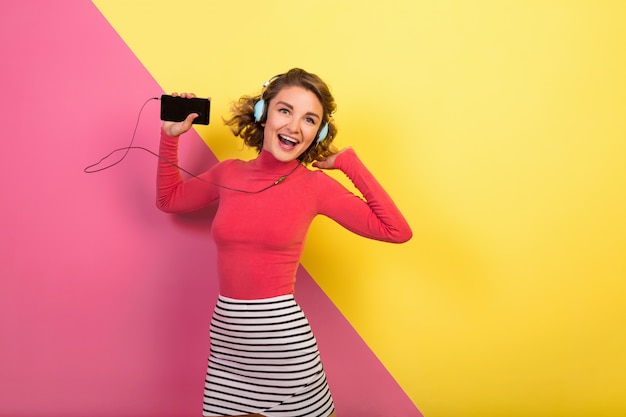 Sonriente mujer atractiva en elegante traje colorido bailando y escuchando música en auriculares