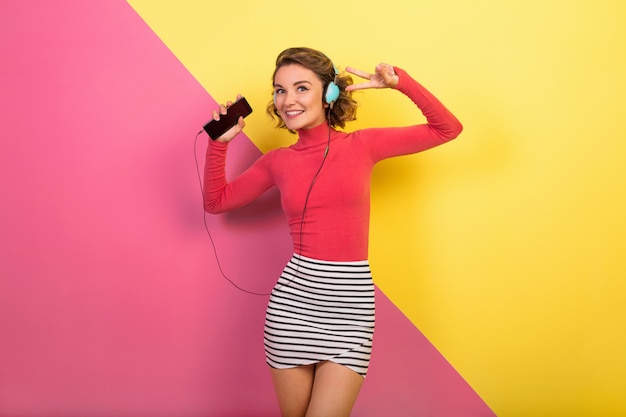 Sonriente mujer atractiva en elegante traje colorido bailando y escuchando música en auriculares