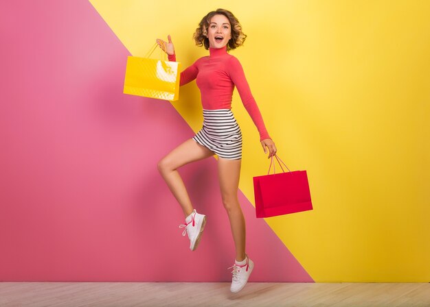 Sonriente mujer atractiva en elegante traje colorido saltando con bolsas de la compra, feliz, fondo amarillo rosa, cuello polo, minifalda rayada, venta, discout, adicta a las compras, tendencia de moda de verano, emocional