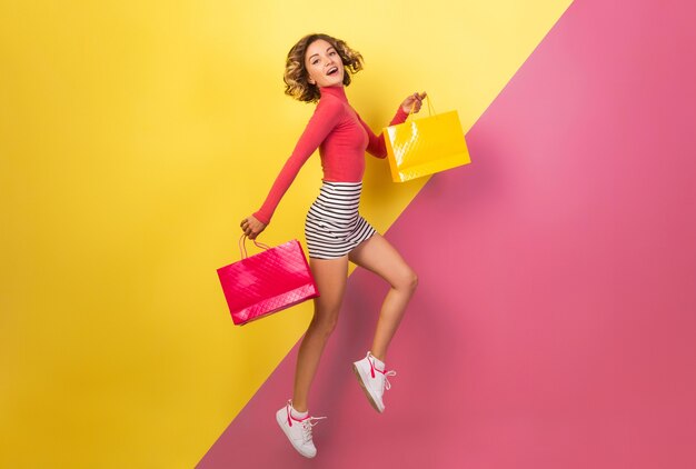 Sonriente mujer atractiva en elegante traje colorido saltando con bolsas de la compra sobre fondo amarillo rosa, cuello polo, minifalda rayada, adicta a las compras en venta, tendencia de moda de verano