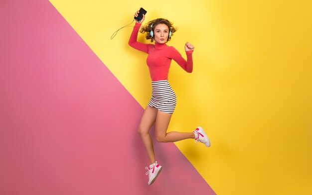 Sonriente mujer atractiva en elegante traje colorido saltando y escuchando música en auriculares