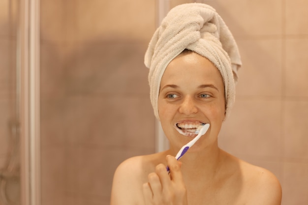 Sonriente mujer atractiva feliz de buen humor cepillándose los dientes, con procedimientos de higiene y belleza en casa en el baño, envuelto en una toalla blanca.