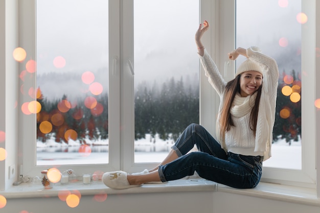 Sonriente mujer atractiva joven feliz en elegante suéter de punto blanco, bufanda y sombrero sentado en casa en el alféizar de la ventana en Navidad divirtiéndose sosteniendo las manos, vista de fondo del bosque de invierno, luces bokeh