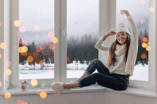 Sonriente mujer atractiva joven feliz en elegante suéter de punto blanco, bufanda y sombrero sentado en casa en el alféizar de la ventana en Navidad divirtiéndose sosteniendo las manos, vista de fondo del bosque de invierno, luces bokeh