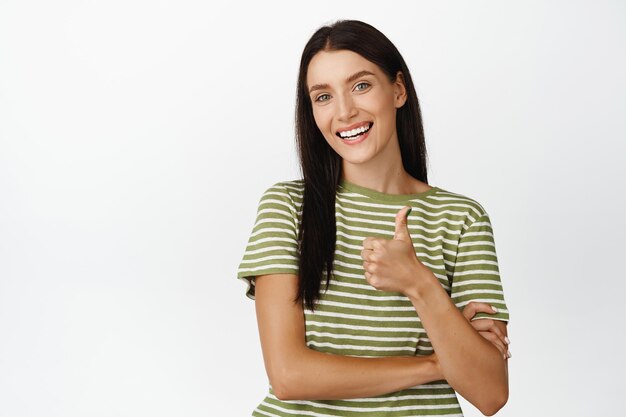 Sonriente mujer atractiva mostrando los pulgares hacia arriba felicitando algo aprobar y como estar de pie sobre fondo blanco.