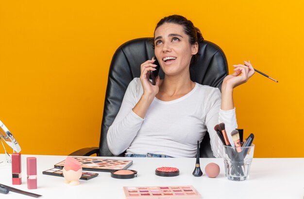 Sonriente mujer bastante caucásica sentada a la mesa con herramientas de maquillaje hablando por teléfono sosteniendo pincel de maquillaje y mirando hacia arriba