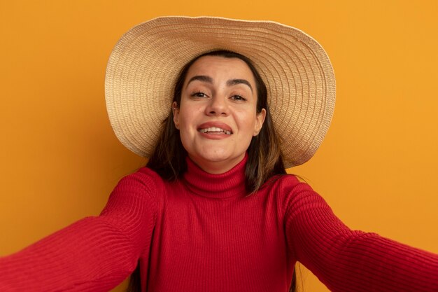 Sonriente mujer bastante caucásica con sombrero de playa pretende sostener la cámara en naranja