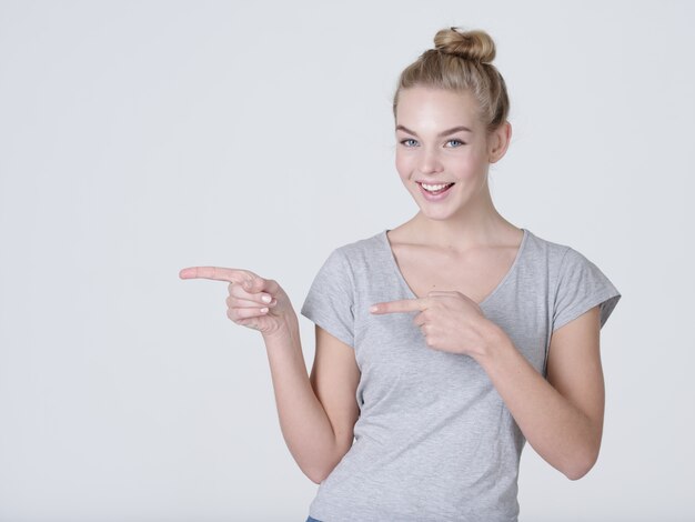 Sonriente mujer bonita apuntando con el dedo sobre fondo gris. Mirando a la camara