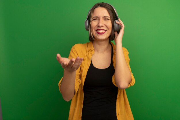 Sonriente mujer bonita en auriculares tiene la mano abierta aislada en la pared verde