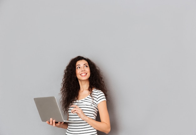 Sonriente mujer bonita en camiseta a rayas con la cara hacia arriba pensando o soñando despierto mientras trabaja a través de la computadora portátil que está aislada sobre la pared gris