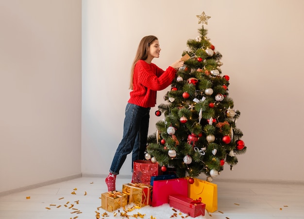 Sonriente mujer bonita emocionada en suéter rojo de pie en casa decorando el árbol de Navidad rodeado de regalos y cajas de regalo