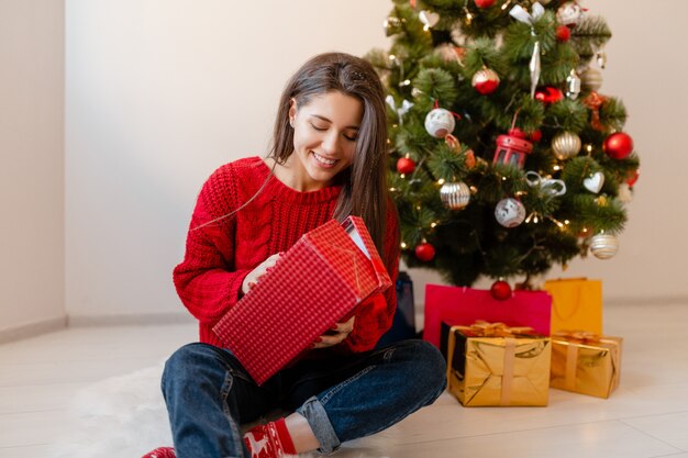 Sonriente mujer bonita emocionada en suéter rojo sentado en casa en el árbol de Navidad desembalaje de regalos y cajas de regalo