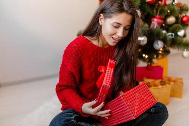 Sonriente mujer bonita emocionada en suéter rojo sentado en casa en el árbol de Navidad desembalaje de regalos y cajas de regalo