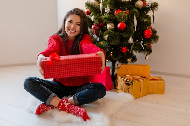 Sonriente mujer bonita emocionada en suéter rojo sentado en casa en el árbol de Navidad desembalaje de regalos y cajas de regalo