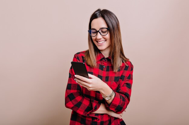 Sonriente mujer bonita lleva gafas de desplazamiento smartphone con emociones felices os pared aislada