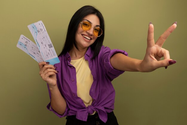 Sonriente mujer bonita morena en gafas de sol gestos signo de la mano de la victoria y tiene boletos de avión aislados en la pared verde oliva