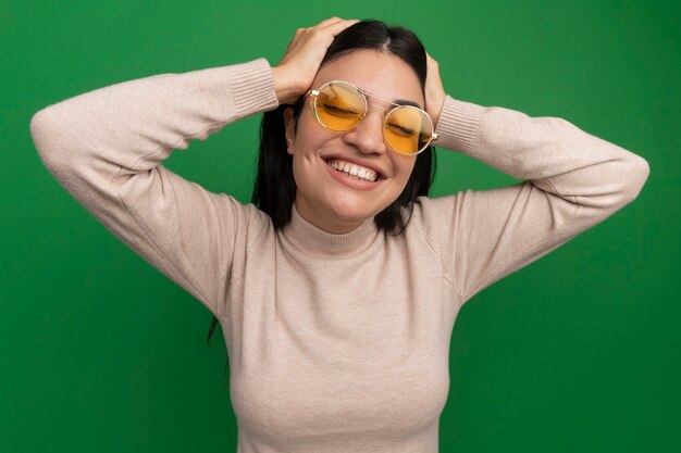 Sonriente mujer bonita morena con gafas de sol pone las manos en la cabeza aislada en la pared verde