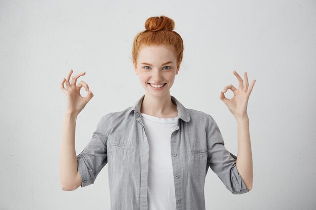 Sonriente mujer bonita con un nudo de pelo rojo con camisa que muestra el signo de Ok con los dedos con una sonrisa sincera disfrutando de su vida con la esperanza de que todo sea genial. Gente y felicidad