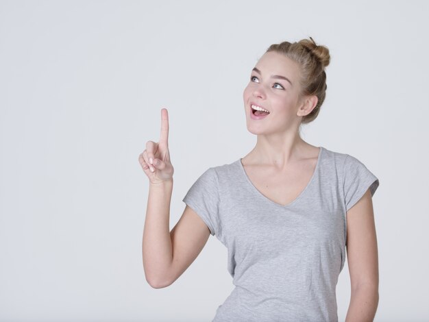 Sonriente mujer bonita que señala el dedo hacia arriba sobre fondo gris. Mirando a la camara