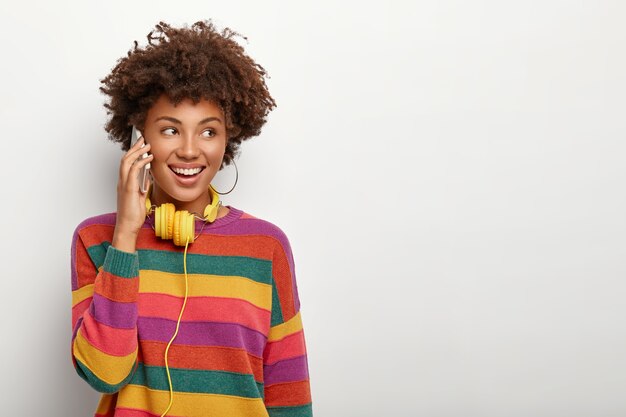 Sonriente mujer bonita tiene conversación telefónica, está de muy buen humor durante una conversación agradable, sostiene el teléfono inteligente cerca de la oreja, usa un suéter informal, usa auriculares, aislado en blanco