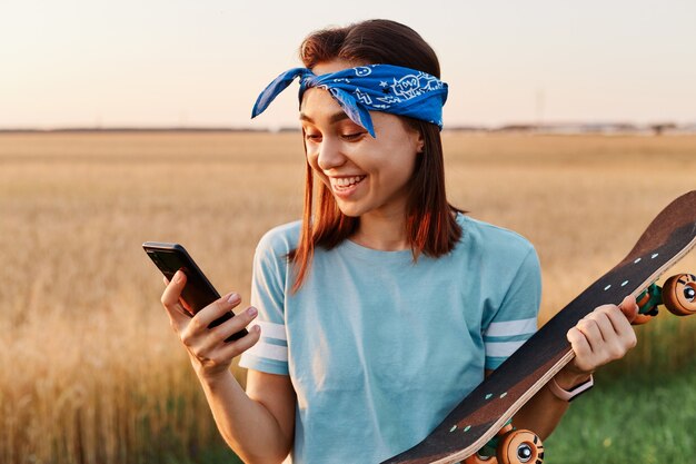 Sonriente mujer con cabello oscuro vistiendo t mierda y banda para el cabello sosteniendo en las manos patineta y teléfono inteligente, mirando la pantalla del teléfono celular con felicidad, leyendo excelentes noticias del mensaje.