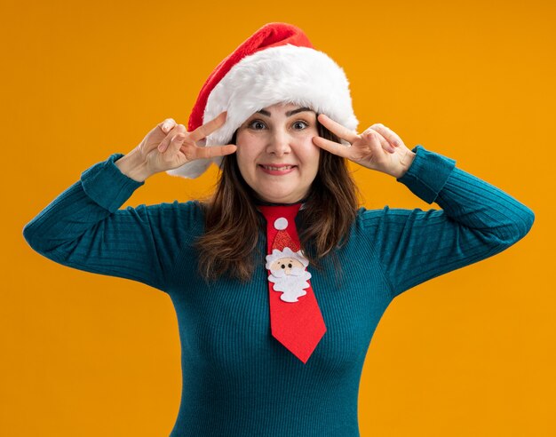 Sonriente mujer caucásica adulta con gorro de Papá Noel y corbata de santa gesticulando el signo de la victoria a través de los dedos aislados en la pared naranja con espacio de copia