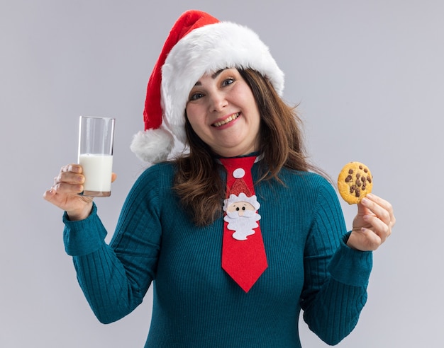 Sonriente mujer caucásica adulta con gorro de Papá Noel y corbata de santa sostiene un vaso de leche y galletas aisladas sobre fondo blanco con espacio de copia