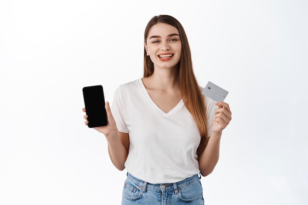 Sonriente mujer caucásica mostrando tarjeta de crédito y pantalla de teléfono inteligente, recomendando aplicaciones móviles, tienda de compras en línea, página web en la pantalla del teléfono, de pie sobre fondo blanco.