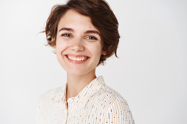 Sonriente mujer caucásica con pelo corto, mirando feliz de pie en blusa contra la pared blanca