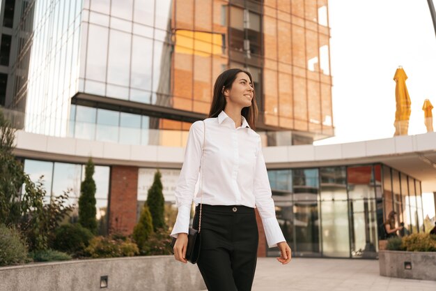 Sonriente mujer caucásica en ropa de negocios caminando cerca del centro de negocios urbano