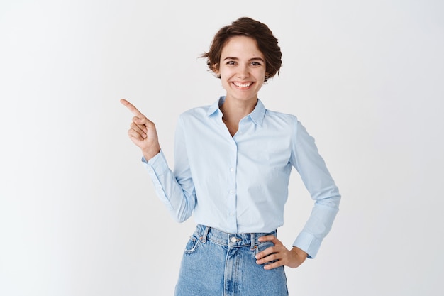 Sonriente mujer ceo profesional con camisa de cuello azul, apuntando con el dedo hacia la izquierda y mirando confiada, de pie sobre la pared blanca